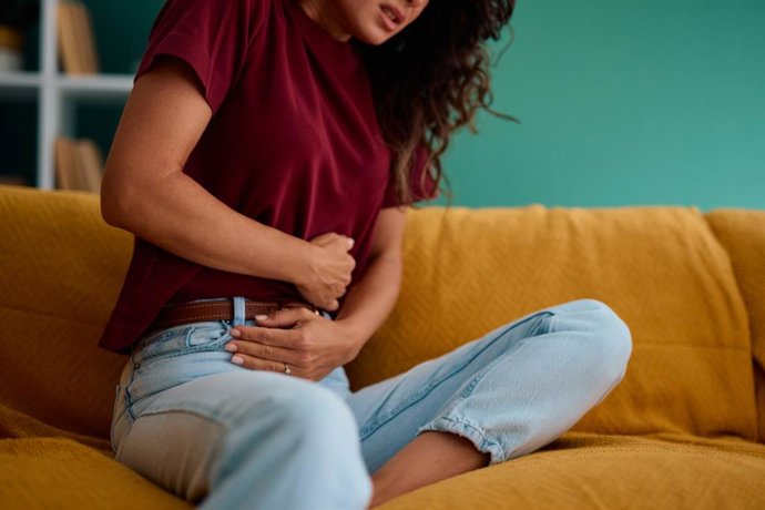 Archivo - Woman experiencing stomach or abdominal pain while sitting on a sofa at home, clutching her abdomen with her hands, indicating discomfort or digestive issues