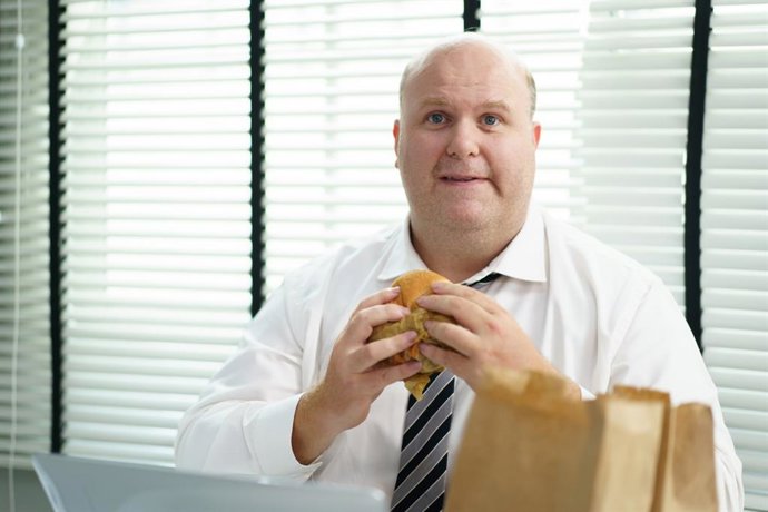 Archivo - Hombre con sobrepeso comiendo hambuerguesa en la oficina, mirando a la c&aacute;mara.